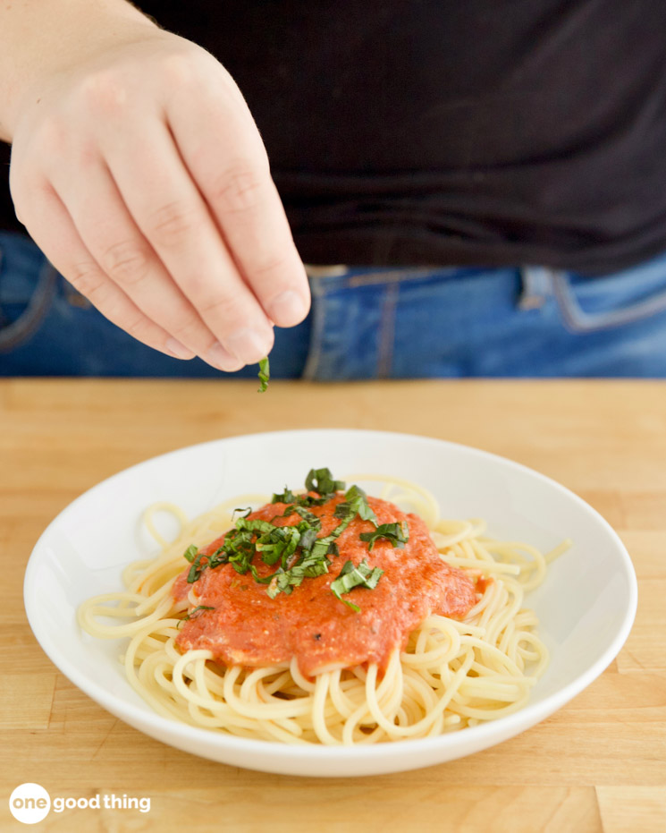 Photo showing a hand sprinkling chopped basil on a plate of spaghetti with pasta sauce.