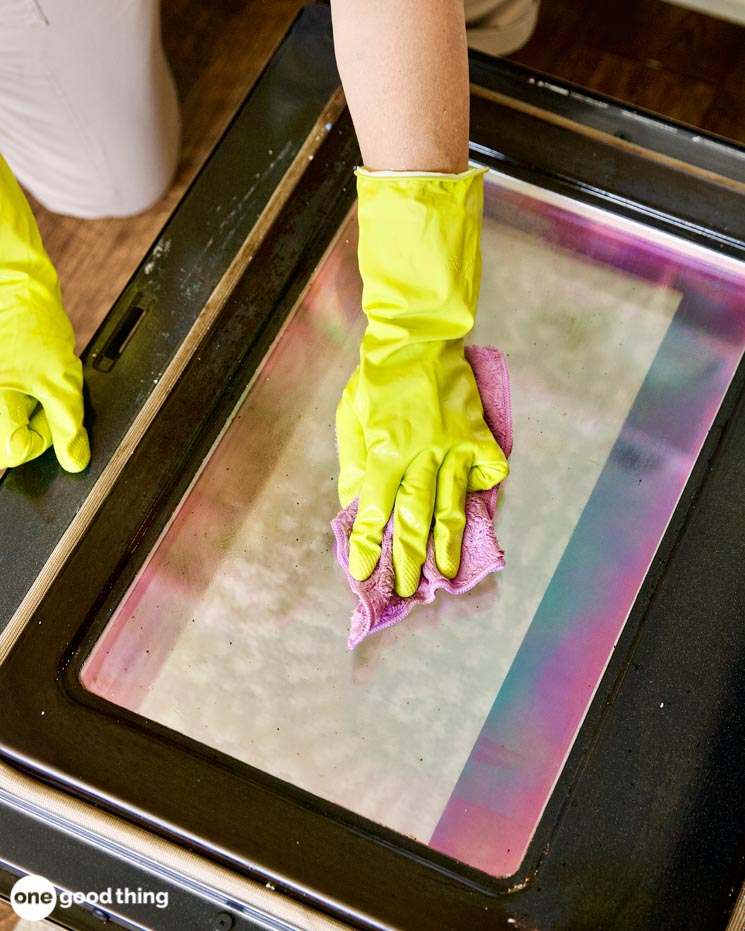 A person in bright yellow gloves diligently cleans the oven door with a vibrant pink cloth, ensuring it sparkles like new.