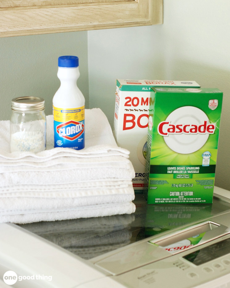 Solving Laundry Problems - stack of bright white towels with a jar of homemade laundry detergent, bottle of bleach, box of borax, and box of dishwasher detergent powder