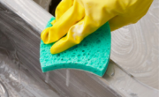 A hand wearing a yellow rubber glove cleans a stainless steel sink using a green sponge.