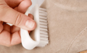 A hand demonstrates how to clean suede shoes by using a small brush with white bristles on a beige suede boot.