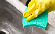 A hand in a yellow glove holding a sponge, scrubbing a stainless steel sink.