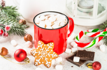 A red mug of indulgent hot chocolate brimming with marshmallows sits amidst a festive spread—snowflake cookie, chocolate pieces, and candy canes—all perfect for cozy winter gatherings, enhanced by ornaments and pine branches. A delightful addition to any hot chocolate bar setup.